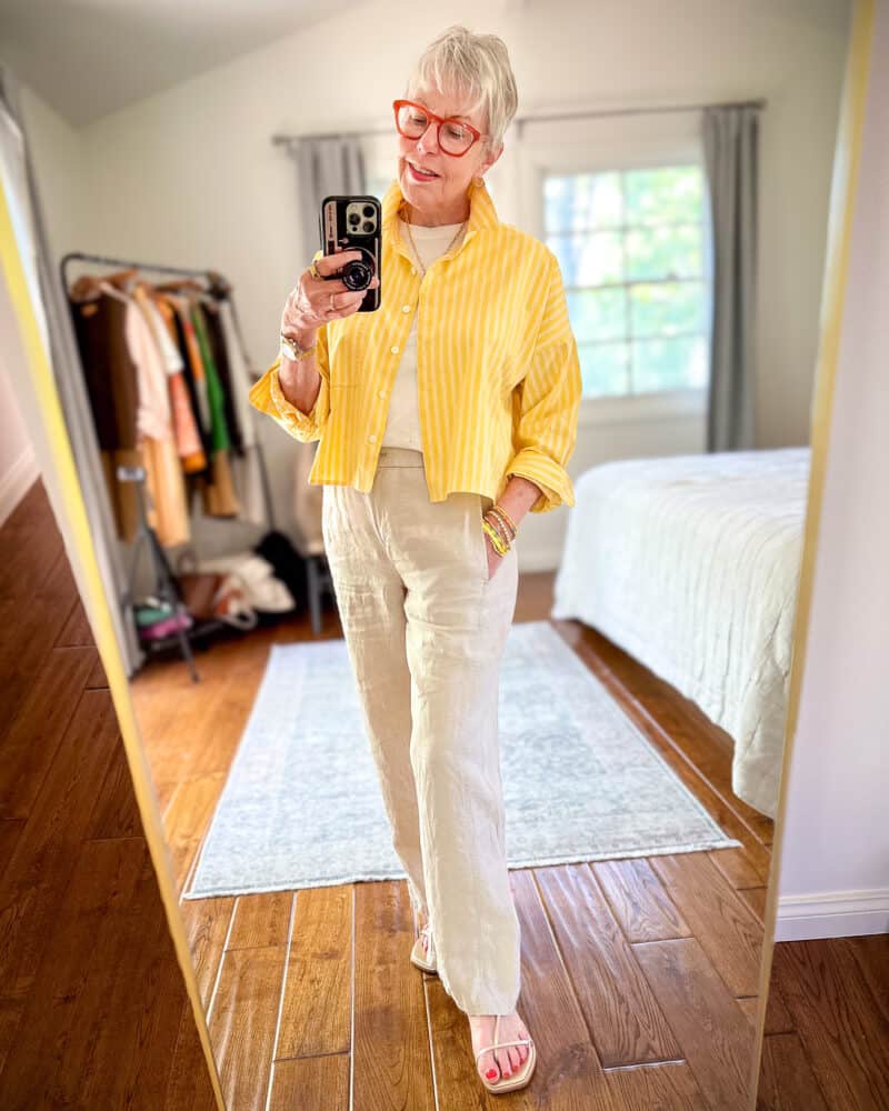 Susan B. wears a yellow striped cropped shirt over a white tee and light beige linen pants, with gold sandals.