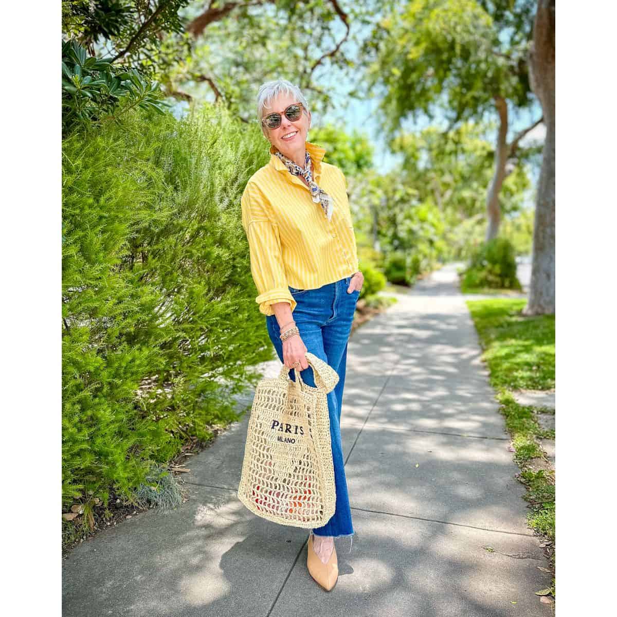 Susan B wears a cropped yellow shirt, patterned bandana, jeans, pointed to flats, carries a straw tote with "Paris/Milano" writing.