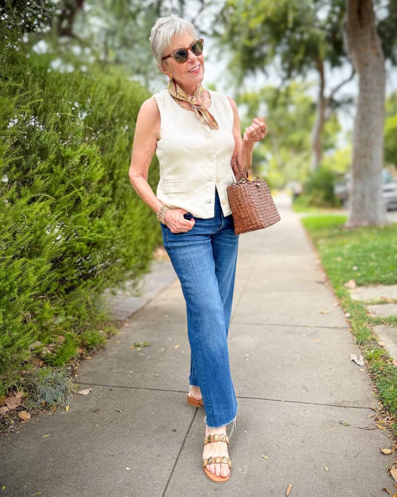 Susan B. wears a linen button-front vest, silk bandana, straight leg jeans, and embellished sandals, carries a woven leather bag.