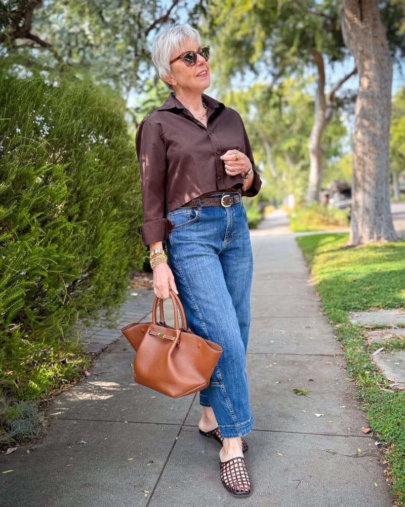 Susan B. wears a brown cropped poplin shirt with a brown belt, barrel-leg jeans, jelly slide sandals, and carries a brown leather bag.