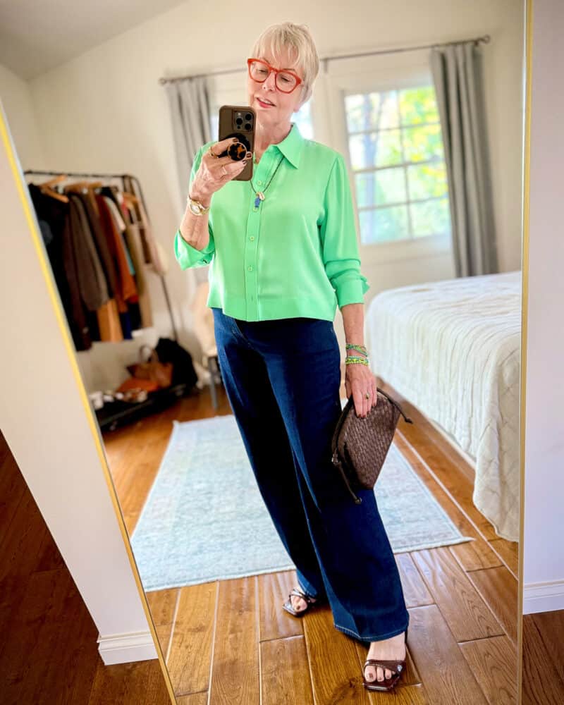 Susan B. wears an apple green silk shirt, dark wash trouser jeans, brown sandals, holds a dark brown woven leather clutch.