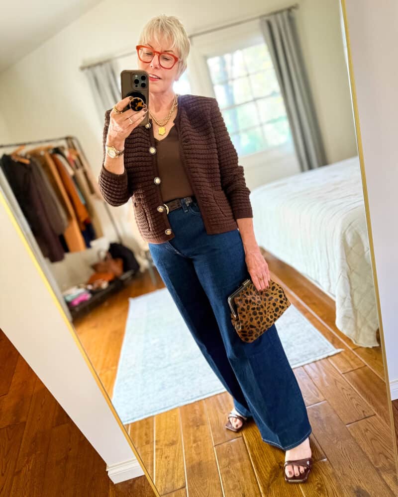 Susan B. wears a textured cardigan with a brown silk top, brown belt, dark wash trouser jeans, brown strappy sandals, carries a leopard print clutch.