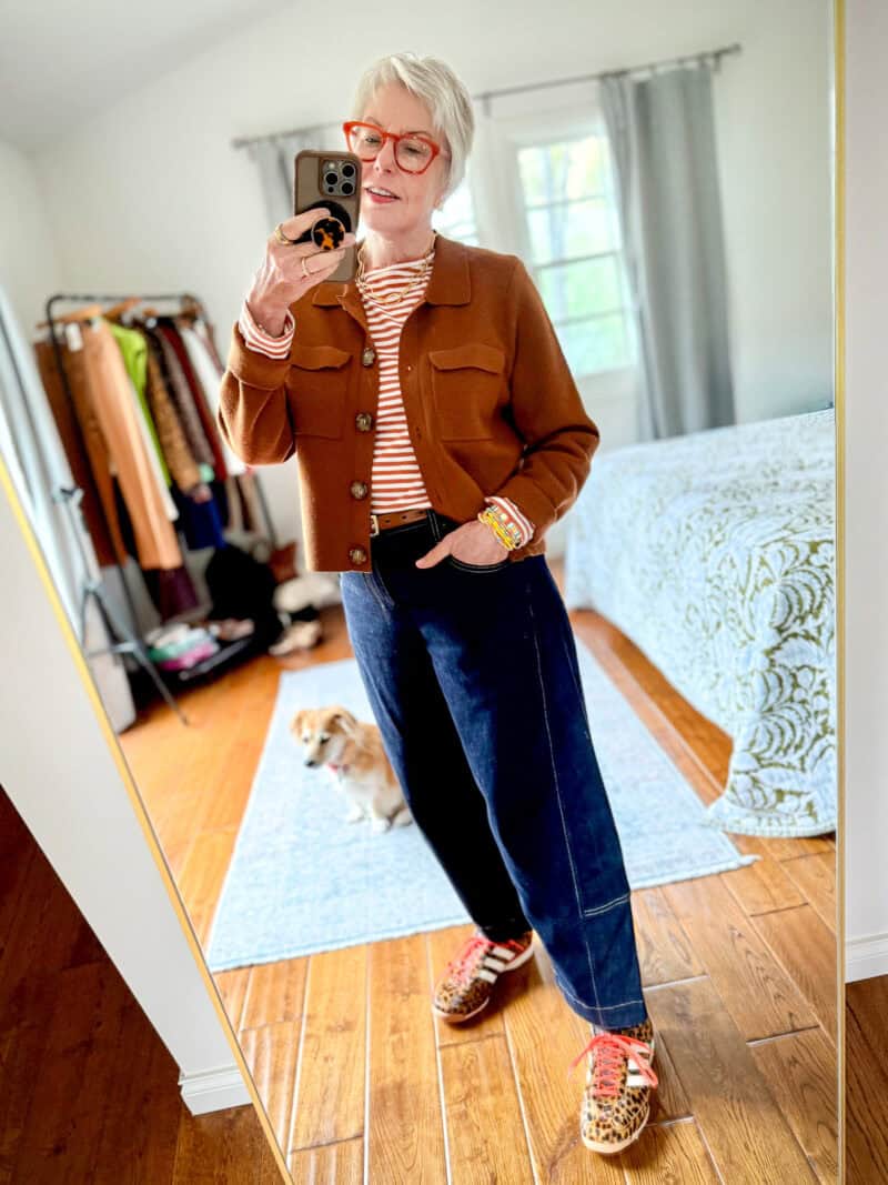 Susan B. wears a brown sweater jacket, striped tee, dark wash barrel leg jeans, and leopard print sneakers.