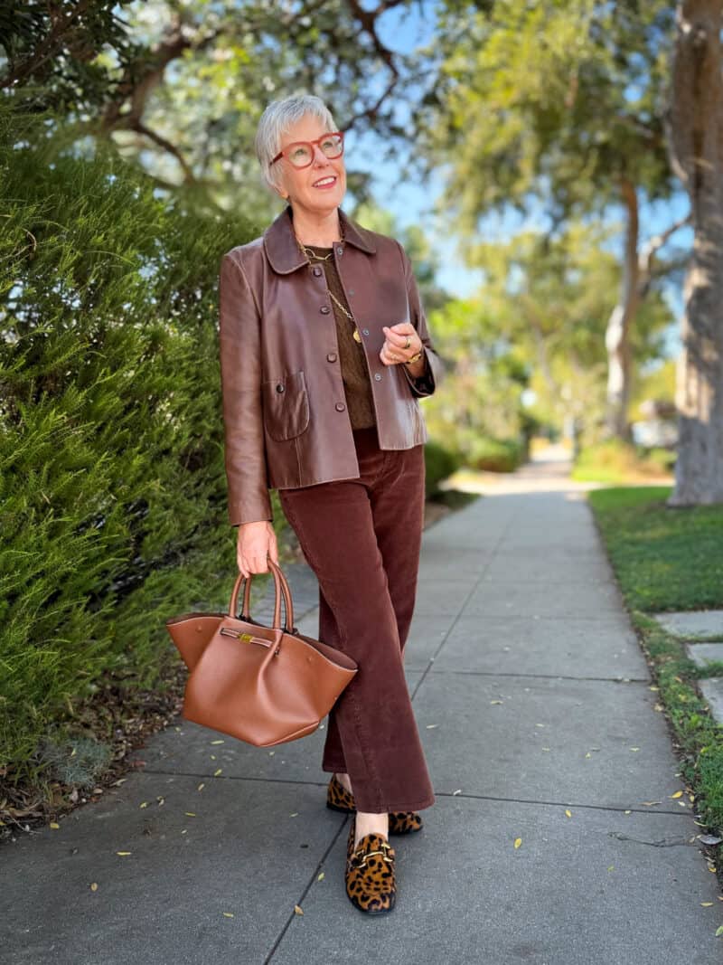 Susan B. wears a brown leather jacket over a brown sweater vest, brown corduroy pants, leopard print loafers, carries a brown leather bag.
