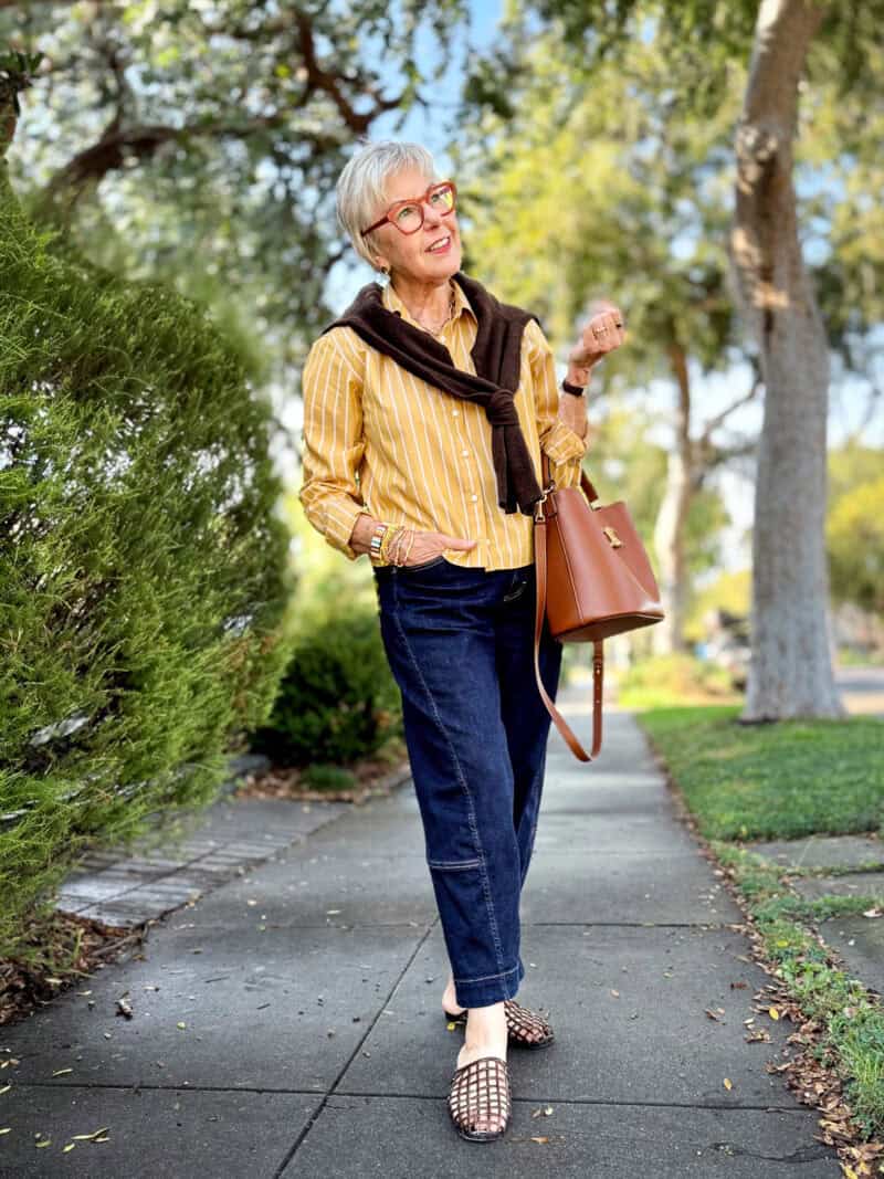 Susan B. wears a honey yellow striped shirt with a dark brown cardigan tied over the shoulders, dark wash barrel jeans, brown jelly slides, carries a brown leather Celine Triomphe bucket bag.