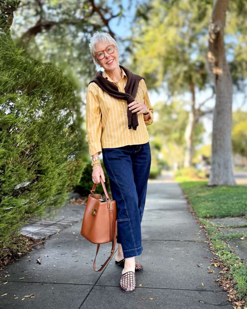 Susan B. wears a honey yellow striped shirt with a dark brown cardigan tied over the shoulders, dark wash barrel jeans, brown jelly slides, carries a brown leather Celine Triomphe bucket bag.