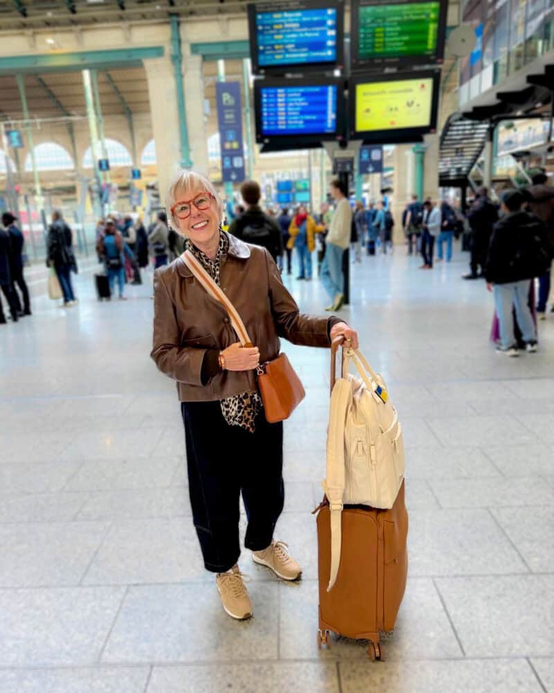 Susan B. arrives in Paris wearing a brown leather jacket, barrel-leg jeans and sneakers. Luggage is spinner bag by Lipaul and Beis backpack.