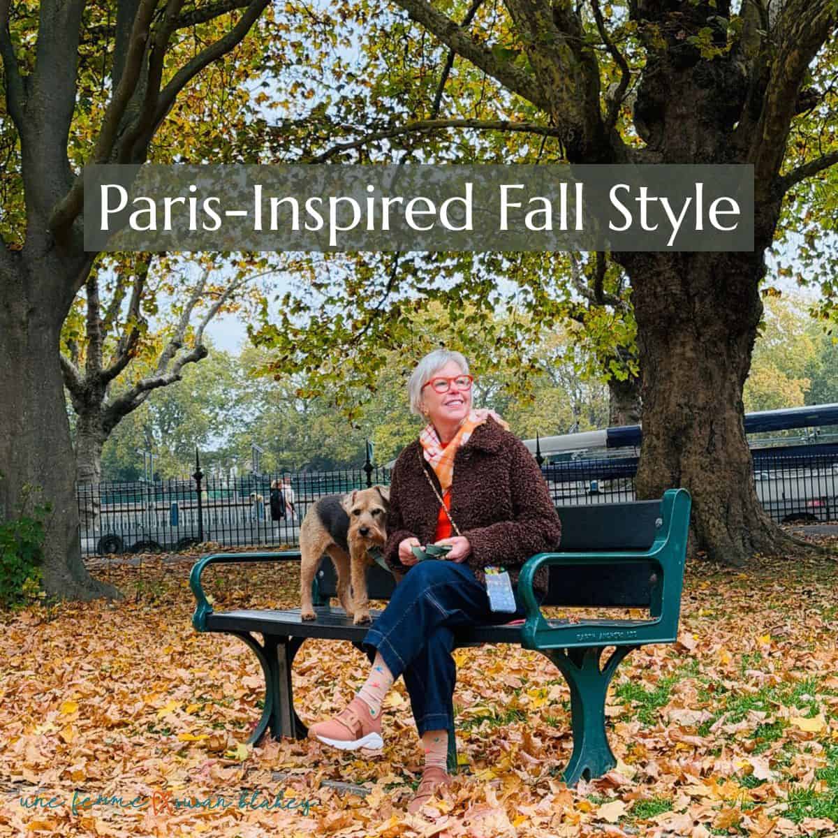 Susan B. sits on a park wearing a faux fur jacket, plaid scarf, red sweater, jeans and sneakers. A terrier dog stands on the bench next to her.