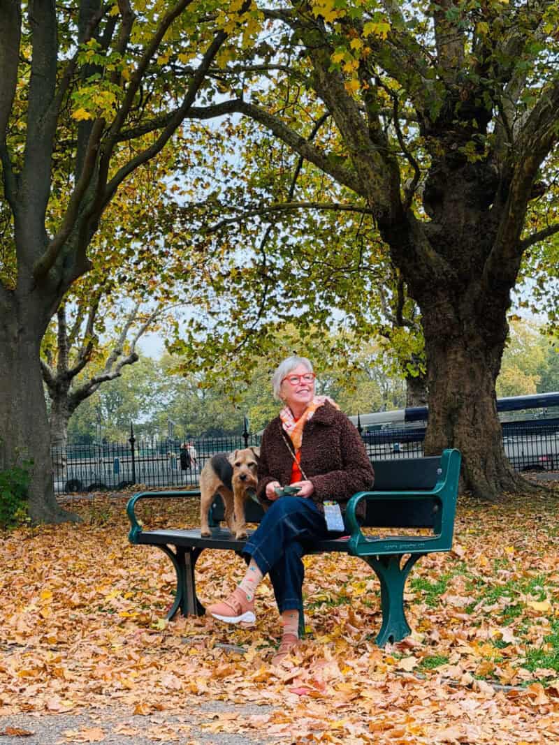Susan B. sits on a park wearing a faux fur jacket, plaid scarf, red sweater, jeans and sneakers. A terrier dog stands on the bench next to her.
