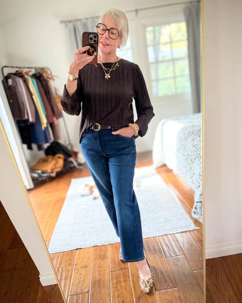 Susan B. wears a dark brown pleated top with layered gold necklaces, dark brown belt, straight leg jeans, leopard print flats.