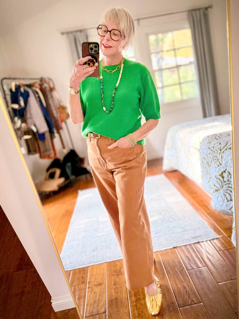 Susan B. wears a green short sleeve cashmere sweater, long pearl and stone necklace, wide leg colored denim jeans, gold ballet flats.