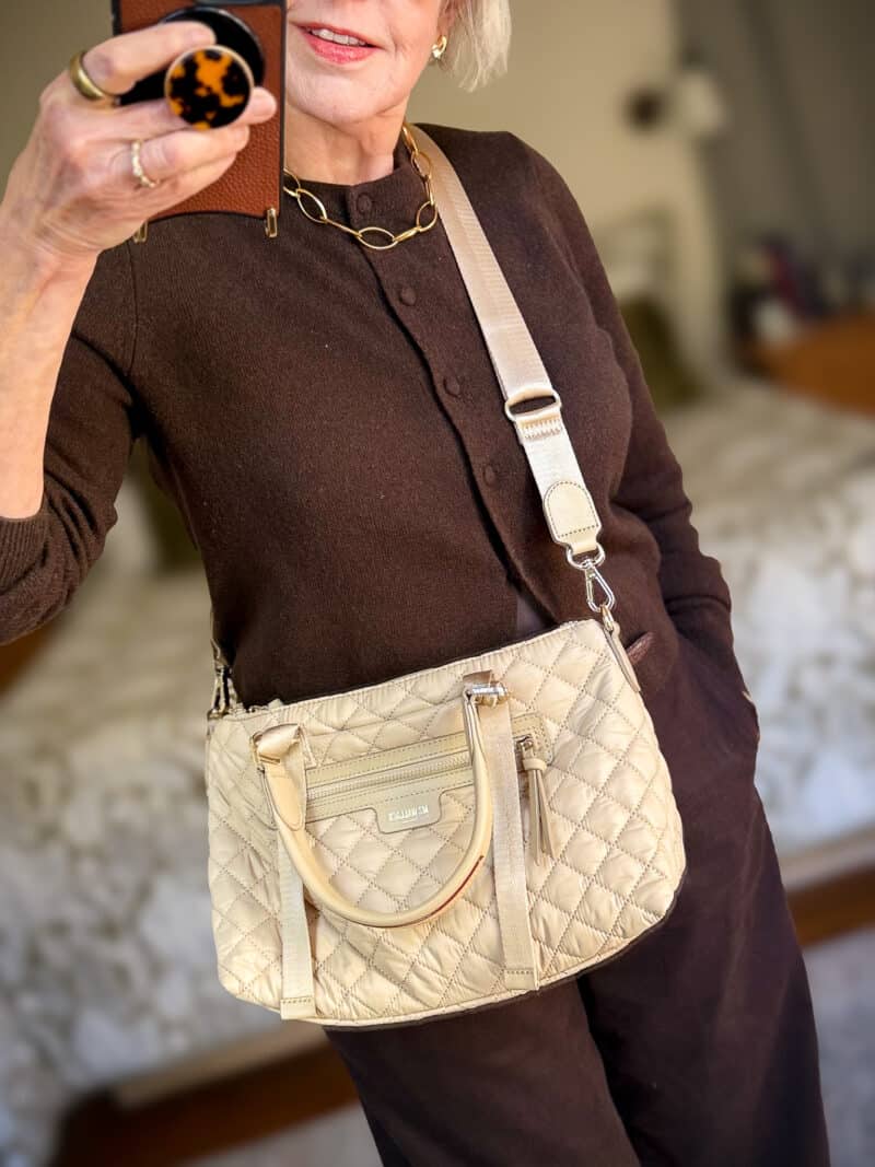 Detail: Susan B. wears MZ Wallace small Park satchel in Buff with crossbody strap over a brown cardigan & trousers.