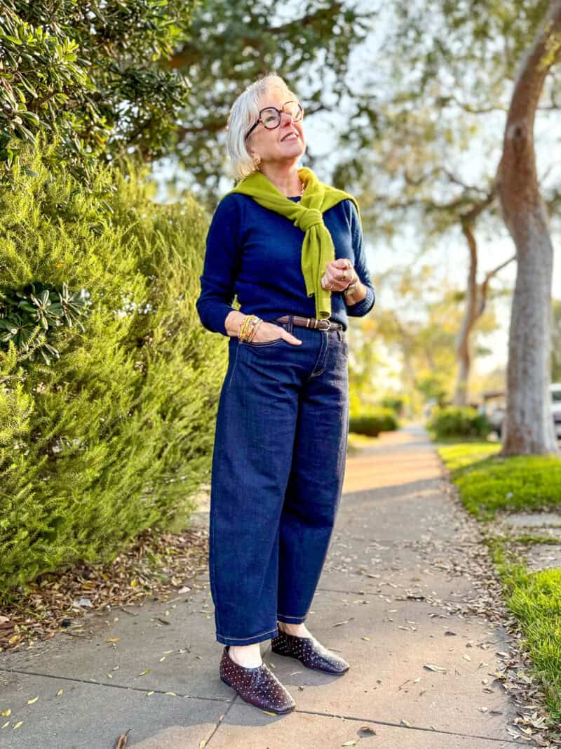Susan B. wears a green cardigan tied over the shoulders, a navy sweater, dark wash barrel-leg jeans and dark brown studded flats.