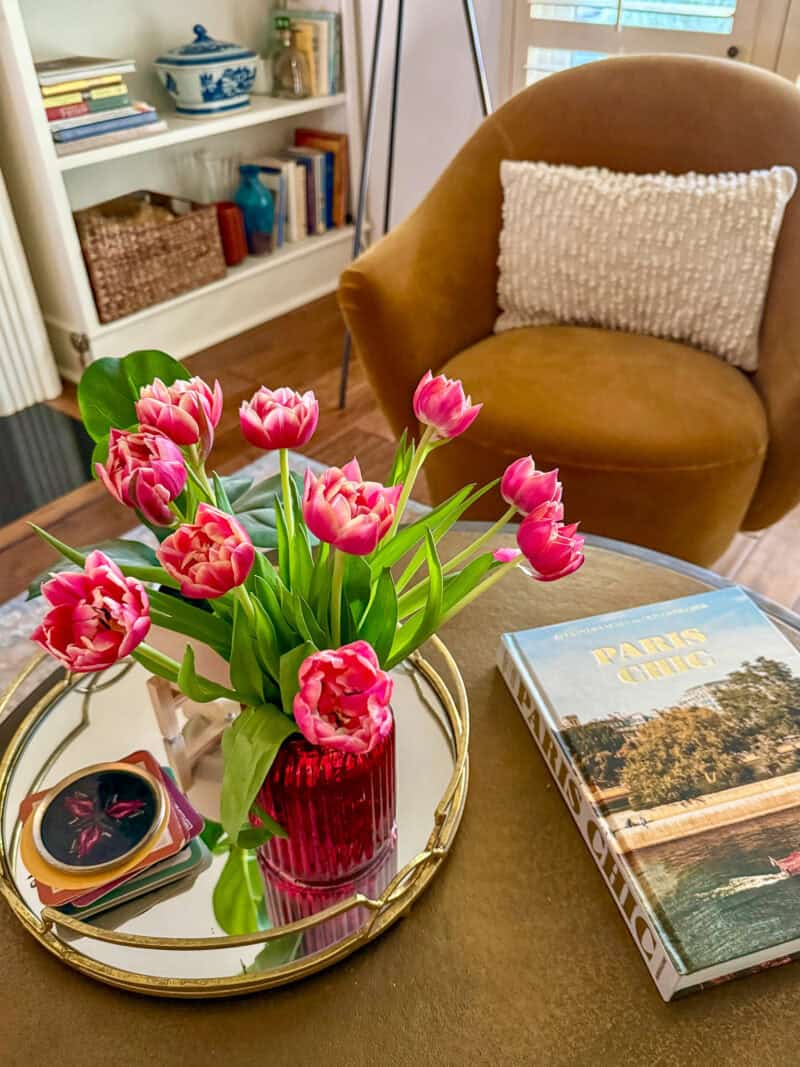 Pink peony tulips in a red vase on a coffee table, with chair and bookshelf in background.