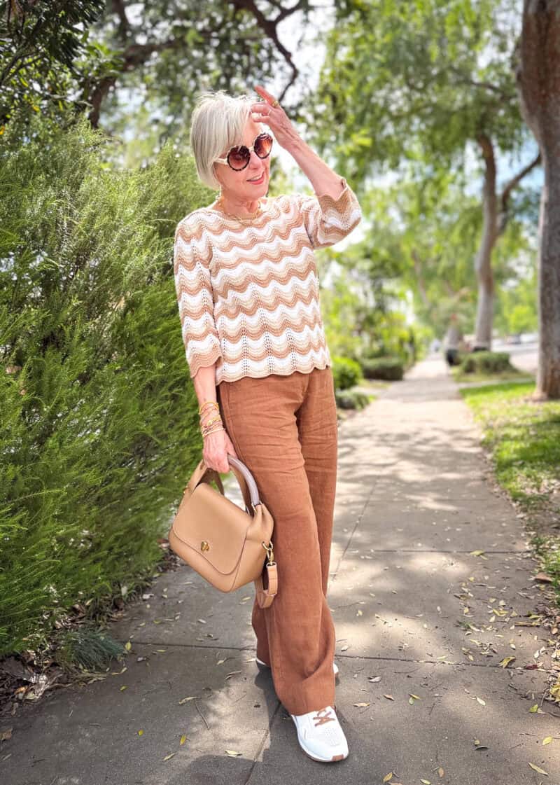 Susan B. wears a striped crochet top, brown linen wide-leg pants, mesh sneakers, and carries a beige leather bag.