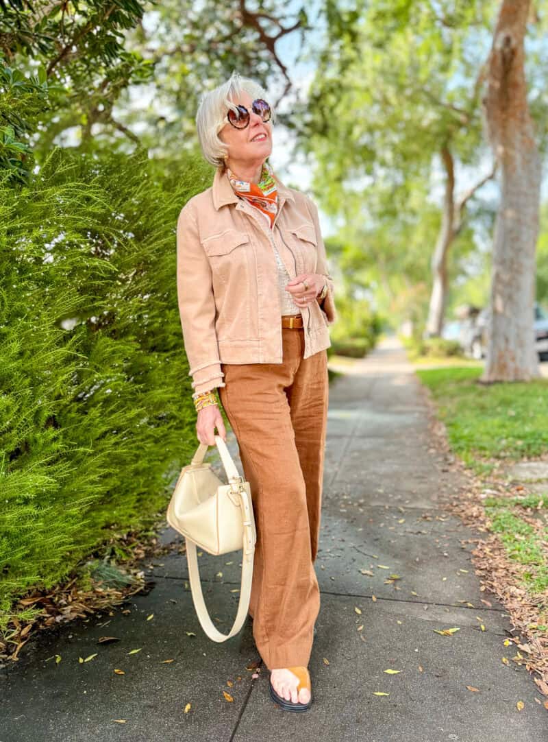 Susan B. wears a tan denim jacket, print bandana, brown linen pants, brown leather sandals, carries a cream leather bag.
