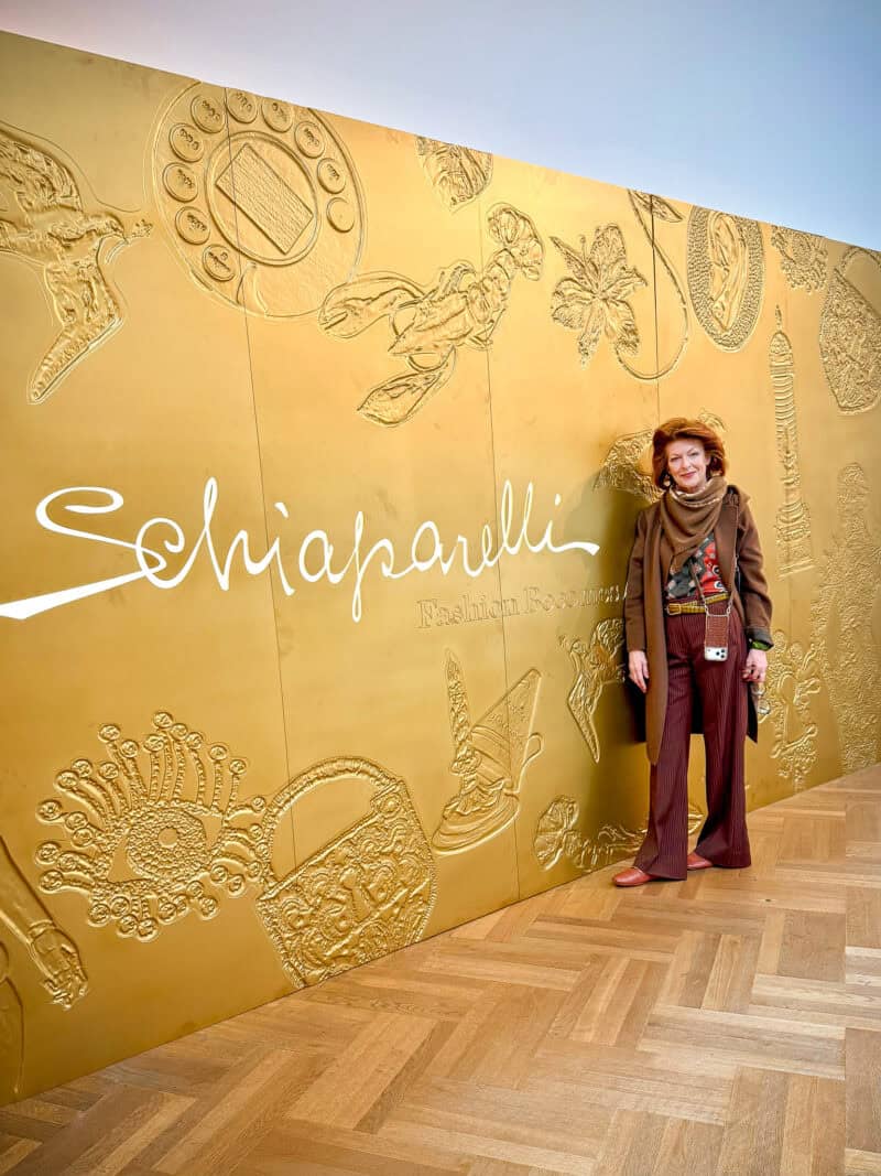 Annie posts by gold Schiaparelli sign at V&A museum, wearing brown and green outfit.