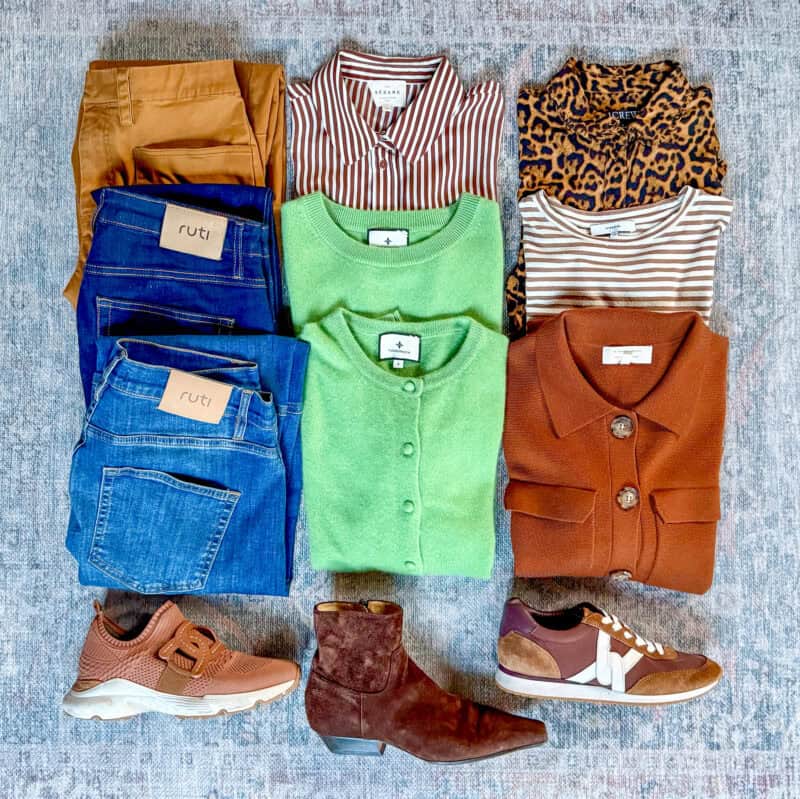 Spring travel capsule wardrobe flatlay arranged on a blue-grey rug. Clothing includes two pairs of Ruti blue jeans, camel-colored trousers, a Sézane brown-and-white striped button-down shirt, a J.Crew leopard-print top, a Vince brown-and-white striped tee, a green button-front cardigan, and a rust-colored knit jacket. Three pairs of shoes are arranged at the bottom: tan Tod's sneakers, brown Veronica Beard lace-up sneakers, and dark brown suede ankle boots.
