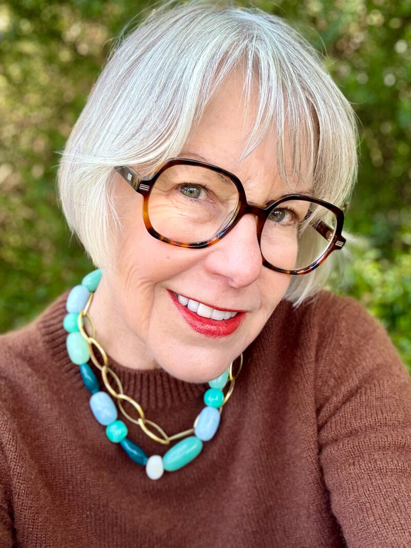 Close-up of a woman with short silver hair, glowing skin, and red lipstick, wearing tortoiseshell glasses and a turquoise necklace outdoors.