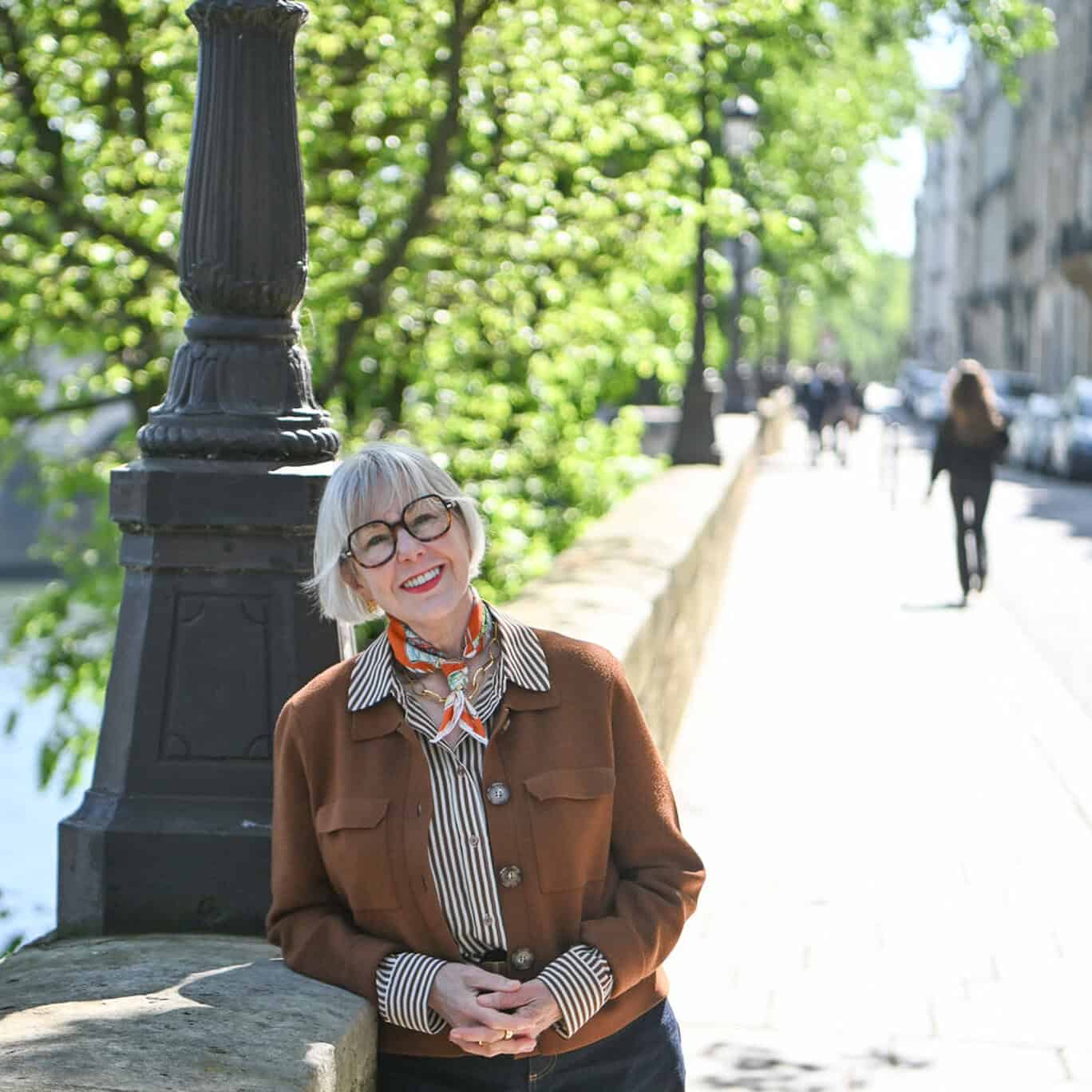 Susan B. leans against a wall along the Seine in Paris wearing a brown jacket, striped shirt, and orange print scarf.