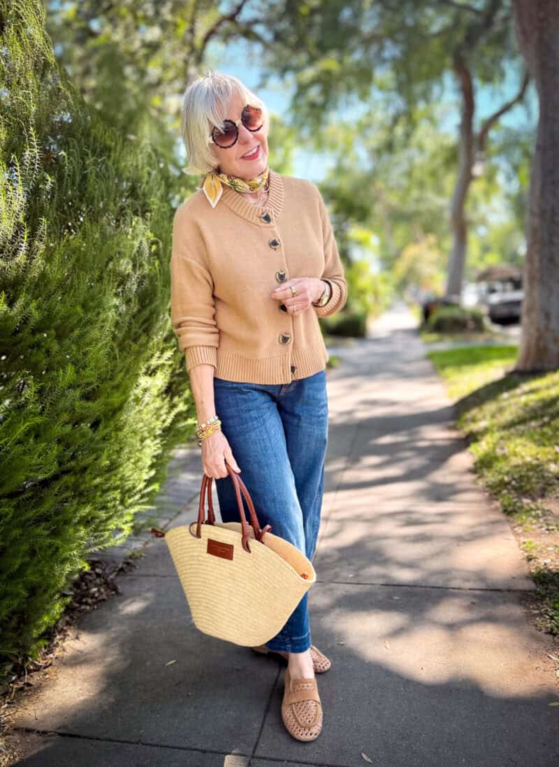 Woman on a sunny sidewalk wearing a camel button-front cardigan, barrel-leg jeans, woven tan loafers, a floral neck scarf, and carrying a woven straw tote bag with cognac leather handles.