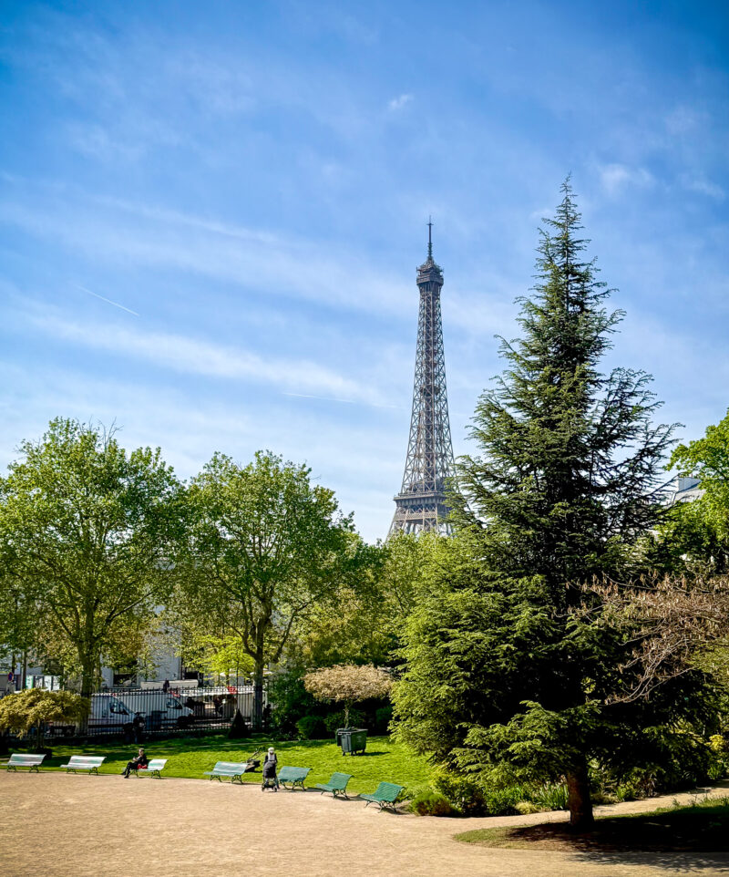 Paris: view of the Eiffel Tower from the Palais Galleria park with green park benches in foreground, people sitting on benches, walking with stroller.
