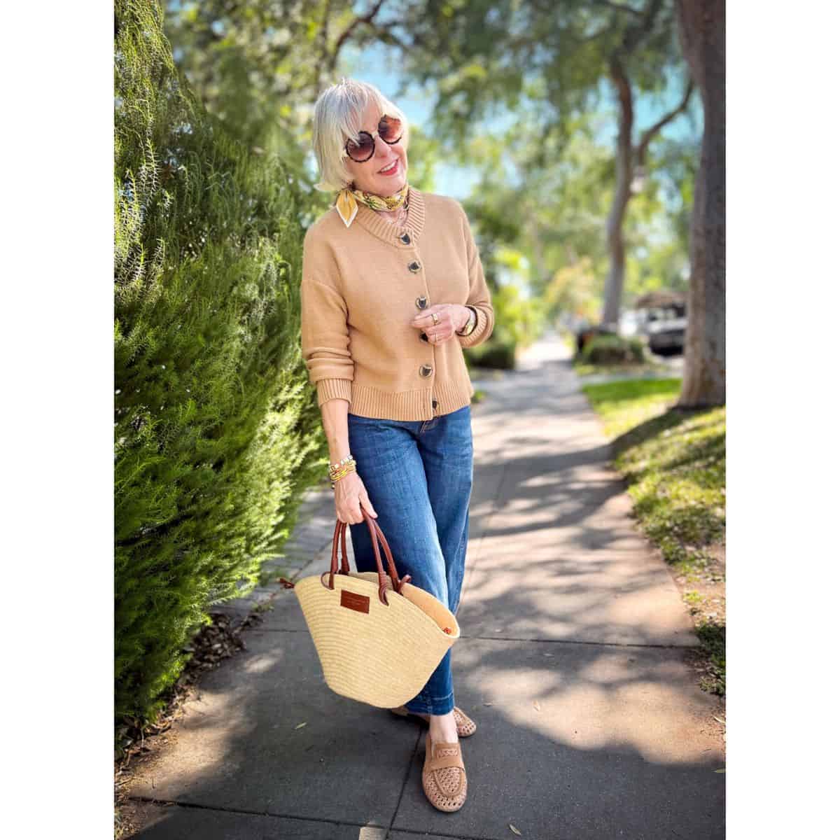 Spring cardigan outfit: Camel button-front cotton cardigan styled with dark wash flare jeans, woven tan loafers, a floral neck scarf, and a woven straw tote bag with cognac leather handles.