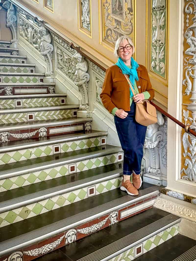 Susan B. stands in a stairwell at the Victoria & Albert museum in London, wearing a turquoise scarf, brown jacket, green sweater, tan bag, blue jeans, and brown sneakers.