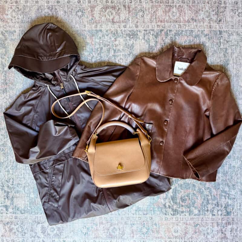 Flat lay of travel outerwear and a handbag on a blue-grey rug. Items include a dark chocolate brown hooded rain anorak, a brown leather snap-front jacket by ba&sh, and a ME+EM tan pebbled leather top-handle crossbody bag with gold hardware.