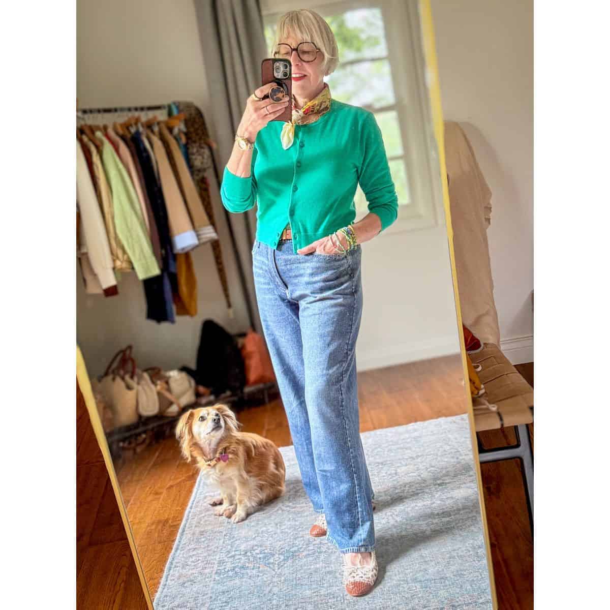 Susan B. takes a mirror selfie while dog Bella looks on. Susan wears a yellow cotton bandana, blue-green cardigan, light wash jeans, colorful bead bracelets, and woven leather cap-toe ballet flats.
