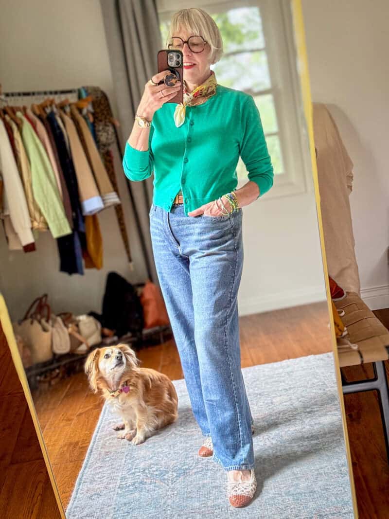 Susan B. takes a mirror selfie while dog Bella looks on. Susan wears a yellow cotton bandana, blue-green cardigan, light wash jeans, colorful bead bracelets, and woven leather cap-toe ballet flats.