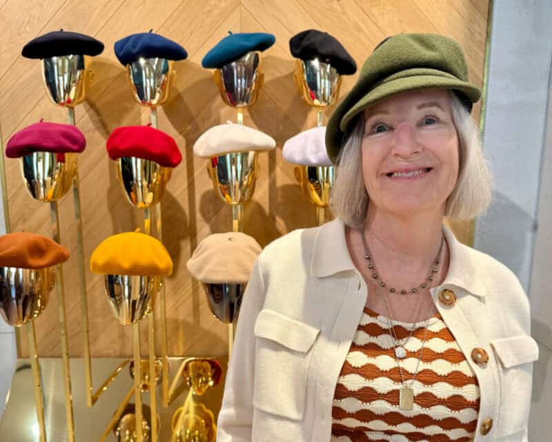 Wall of colorful berets in background, woman in foreground wears an olive newsboy cap with a beige jacket and brown & ivory top.