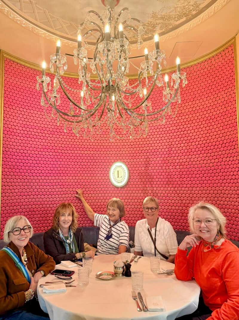 Paris Fashion Tour: five women sit in a booth at Laduree Paris with a wall of pink macarons behind and a crystal chandelier above.