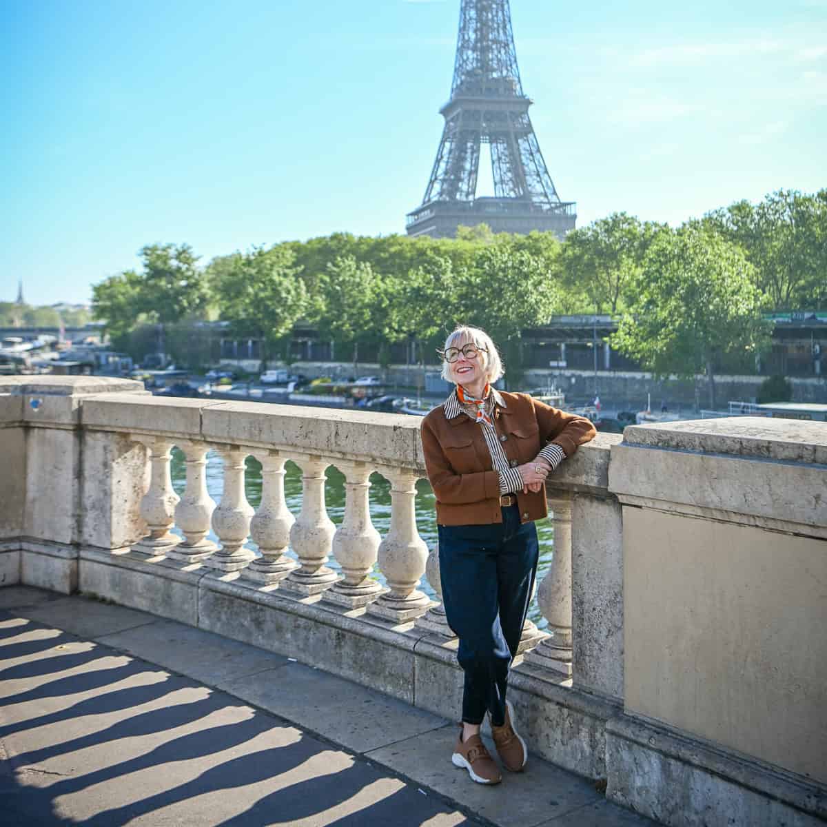 Spring travel outfit on a Paris bridge with the Eiffel Tower in the background — brown knit jacket, striped shirt, navy jeans, silk scarf, and tan sneakers on a sunny day.