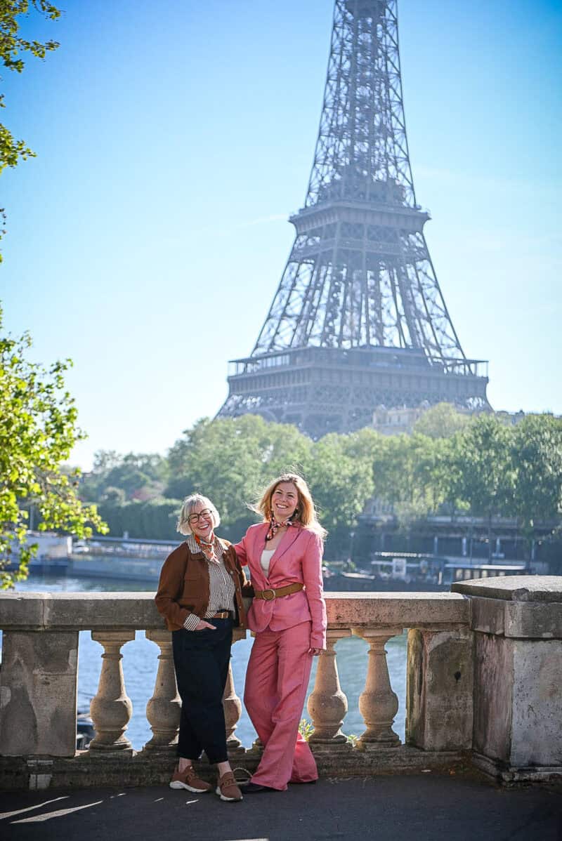 Susan B. and Emma Gely pose on bridge with Eiffel Tower in background. Susan wears a brown jacket, striped shirt & dark wash jeans. Emma wears a pink suit with a tan belt at the waist.