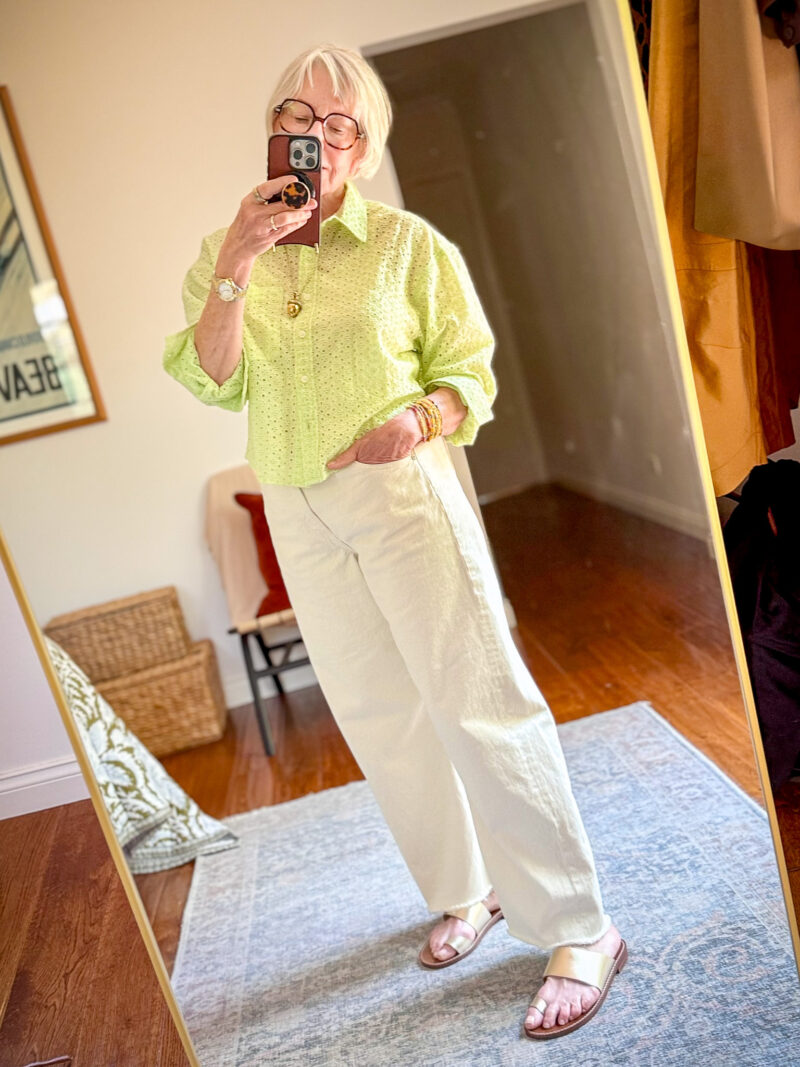 Mirror selfie of a woman wearing a lime green eyelet button-down shirt, wide-leg off-white jeans, and gold flat sandals, accessorized with stacked bracelets, a gold pendant necklace, and tortoiseshell glasses.