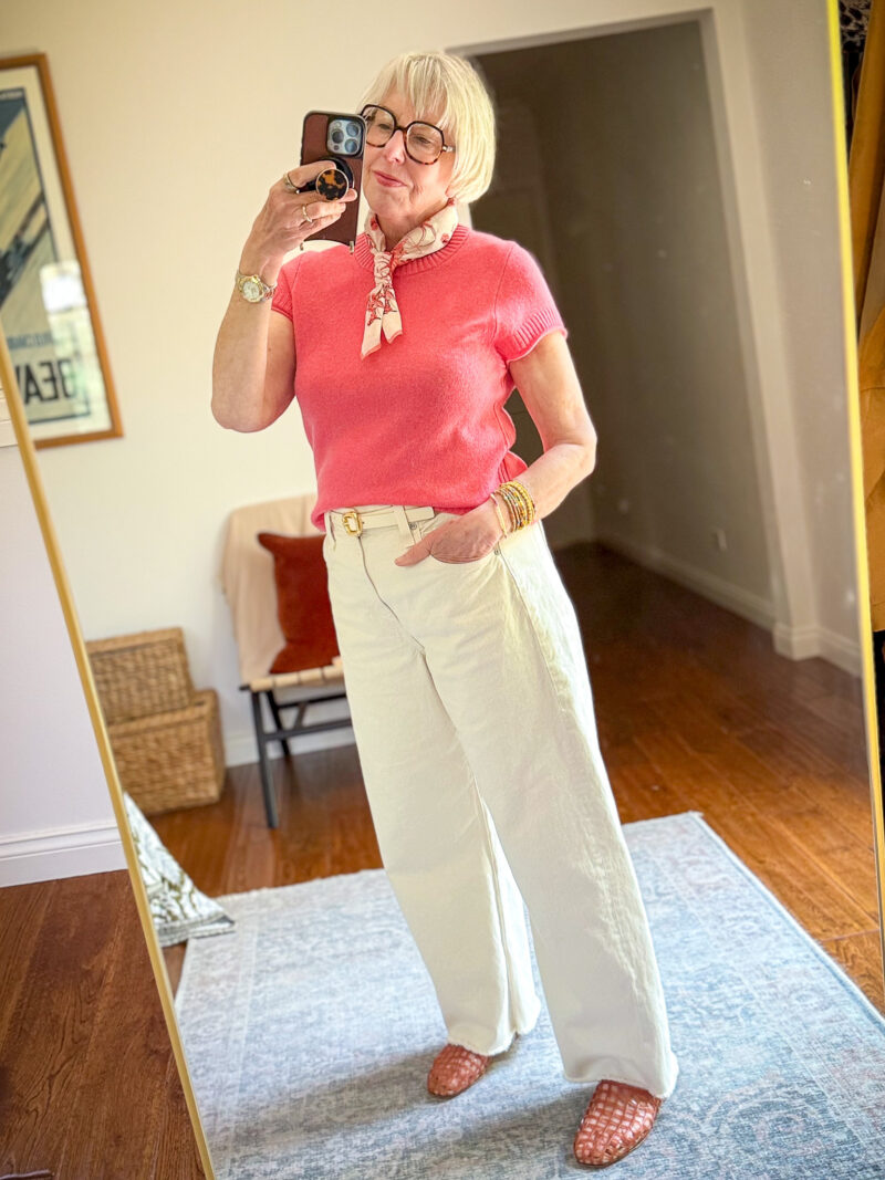 Mirror selfie of a woman wearing a coral pink short-sleeve sweater, wide-leg off-white jeans with a gold-buckle belt, and pink woven flats, styled with a pink printed neck scarf, stacked gold bangles, and tortoiseshell glasses.