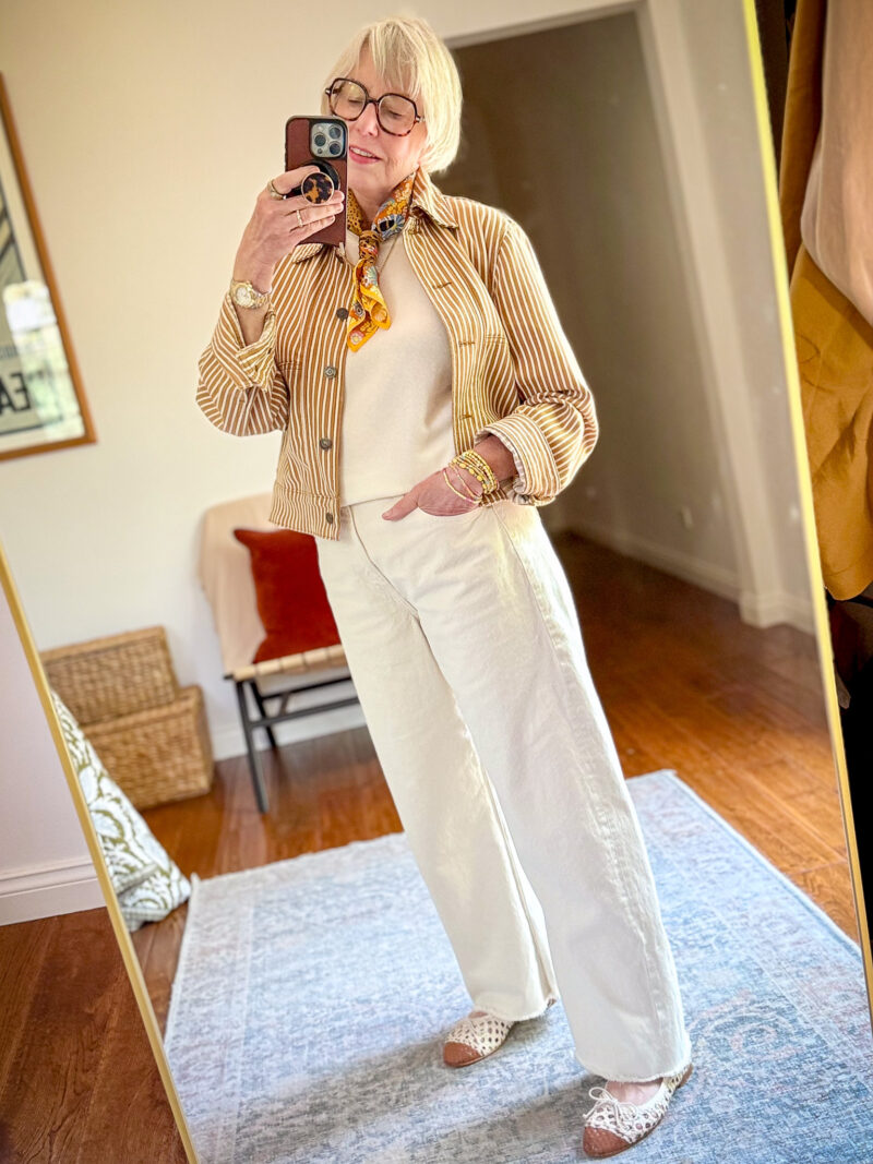 Mirror selfie of a woman wearing a mustard and white striped cropped jacket over a cream top, wide-leg off-white jeans, and white cap-toe open weave ballet flats, accessorized with a gold and orange printed neck scarf, stacked bead bracelets, and tortoiseshell glasses.