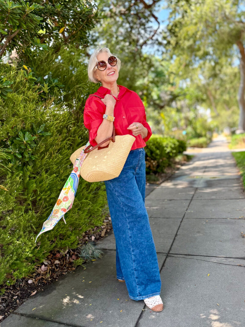 Woman stands on sidewalk wearing a red shirt, blue jeans, and ballet flats, carrying a basket bag with a printed scarf tied to the handle.