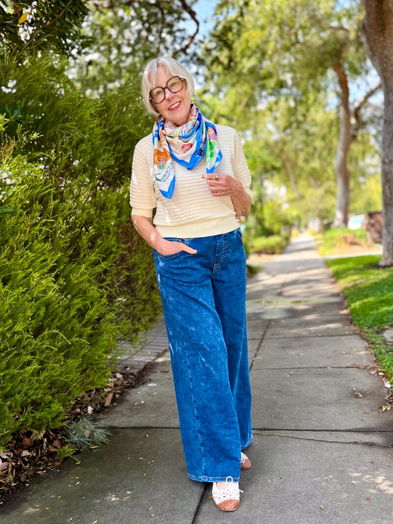 Woman stands on sidewalk wearing a silk square scarf as a kerchief, with an ivory short sleeve sweater, wide leg jeans, and open weave cap-toe ballet flats.
