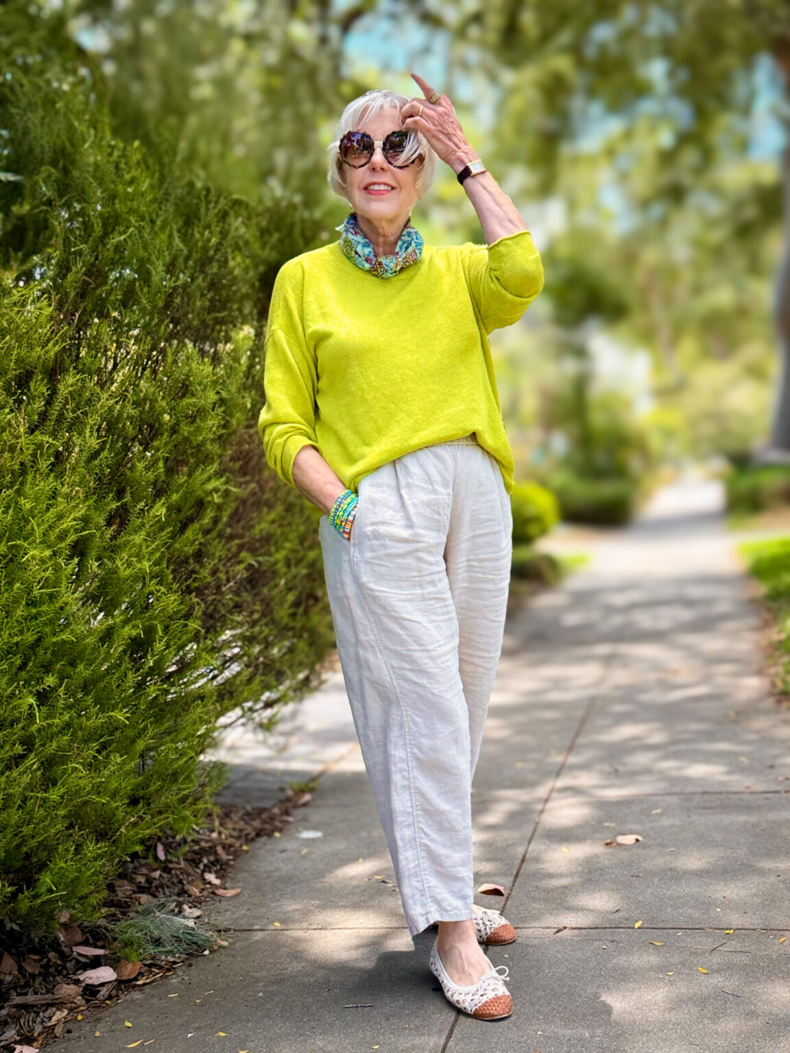 Citron yellow-green linen sweater styled with natural linen trousers, a floral bandana scarf tied at the neck, stacked turquoise bracelets, and white lace-trimmed flats with woven toe detail, worn outdoors on a tree-lined sidewalk
