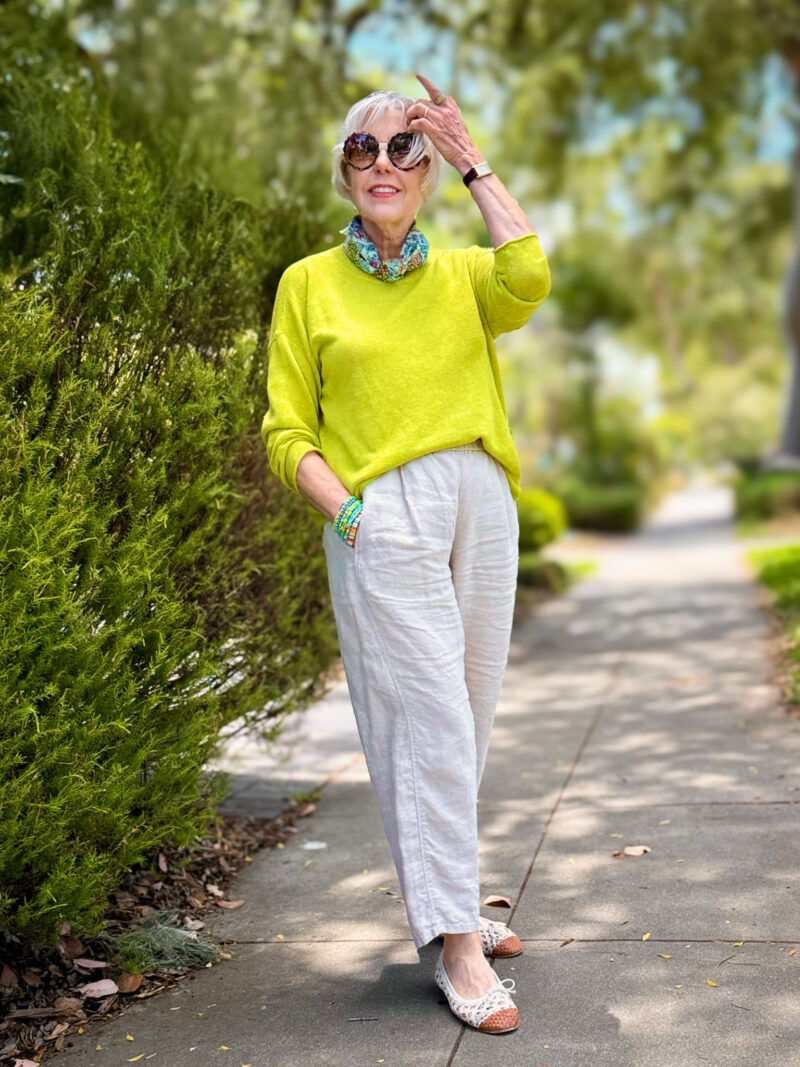 Citron yellow-green linen sweater styled with natural linen trousers, a floral bandana scarf tied at the neck, stacked turquoise bracelets, and white lace-trimmed flats with woven toe detail, worn outdoors on a tree-lined sidewalk