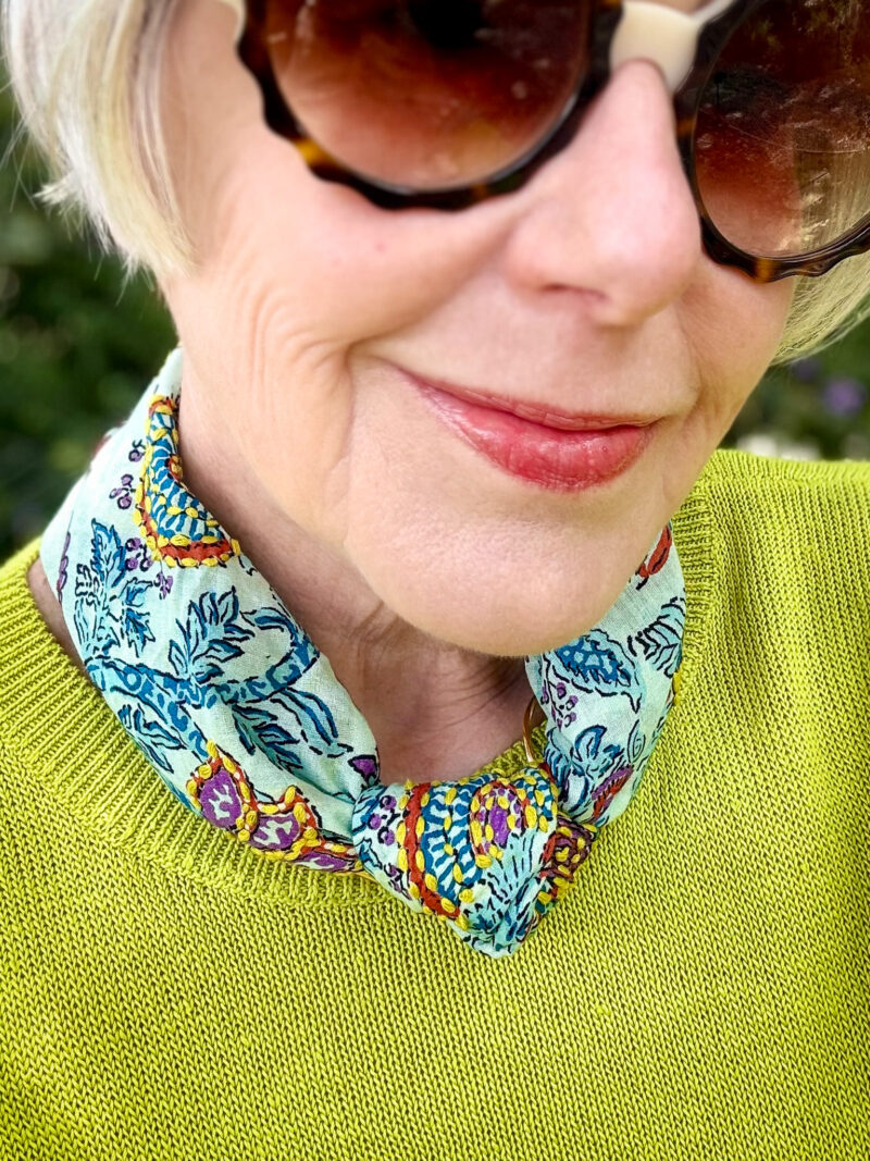 Close-up detail of a block-print bandana scarf in aqua, teal, gold, rust, and purple tied as a necklace at the neckline of a citron linen sweater, with tortoiseshell sunglasses