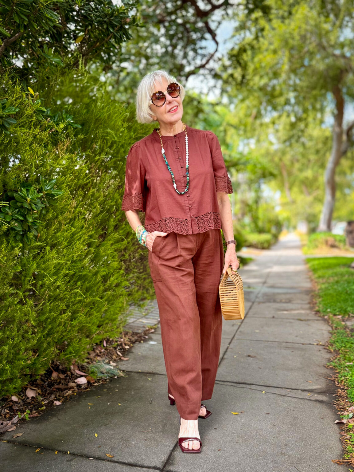 A woman walks along a tree-lined sidewalk wearing a rust-brown outfit — a lace-trimmed short-sleeve top and wide-leg trousers — styled with turquoise beaded necklaces, stacked bracelets, round sunglasses, a bamboo box clutch, and dark brown kitten-heel sandals.