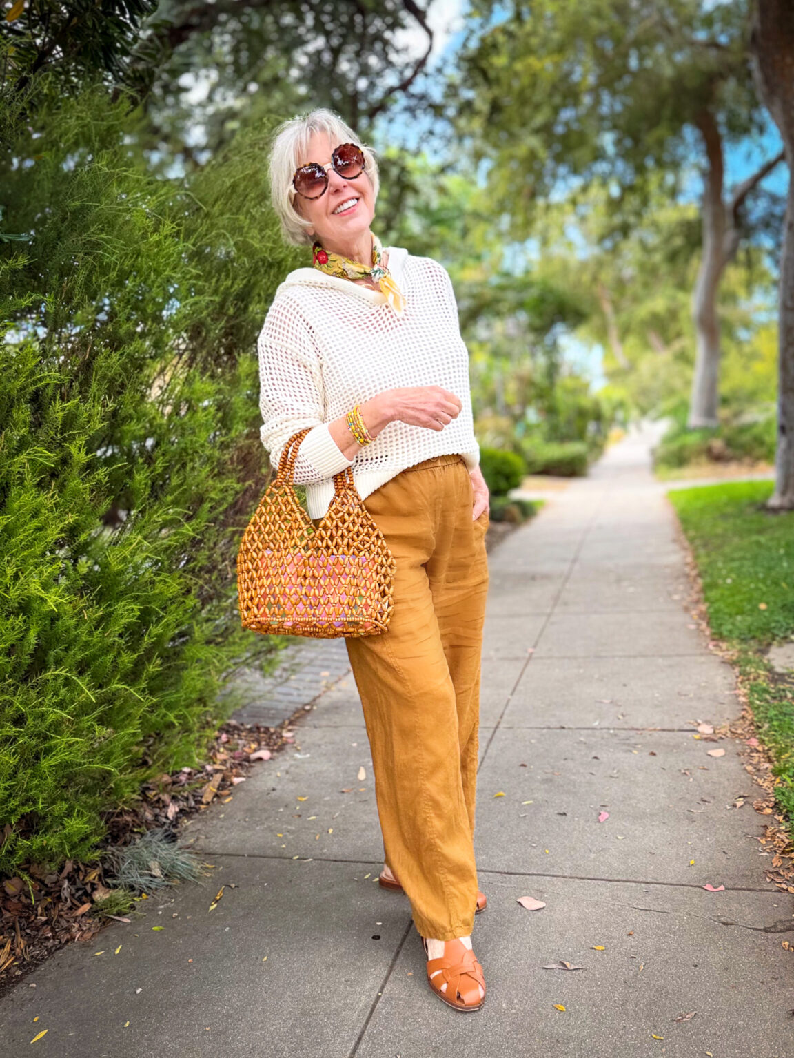 Style blogger wearing a cream open-knit hoodie, golden mustard wide-leg linen pants, floral neck scarf, stacked bracelets, round floral sunglasses, a beaded woven tote in warm orange tones, and tan fisherman sandals, smiling on a sunny suburban sidewalk surrounded by greenery.
