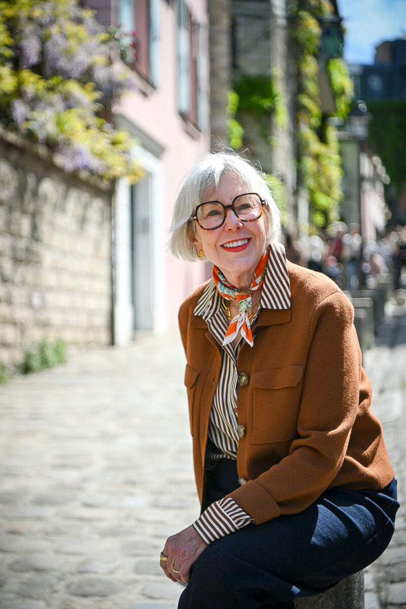 Susan B. sits on a post on a street in Montmartre Paris, wearing an orange cotton bandana, brown striped shirt, brown jacket.