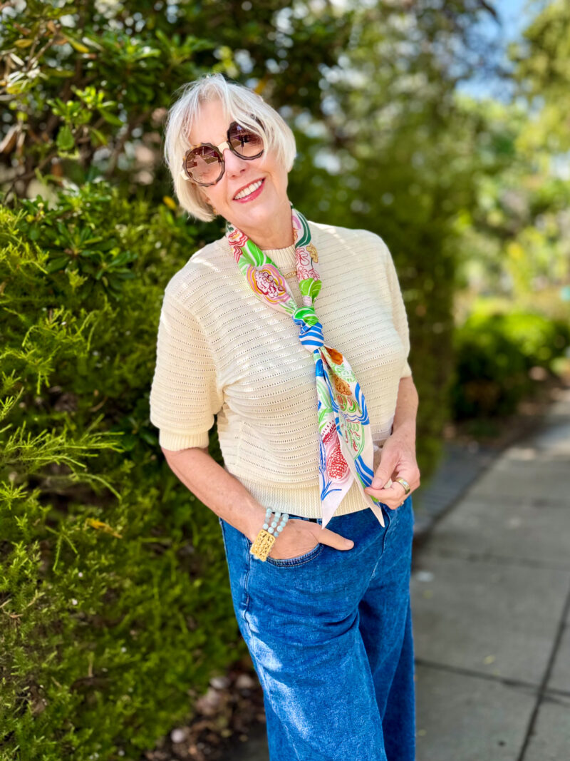 Woman stands on sidewalk wearing silk skinny scarf tied in Windsor knot with textured sweater and blue jeans.