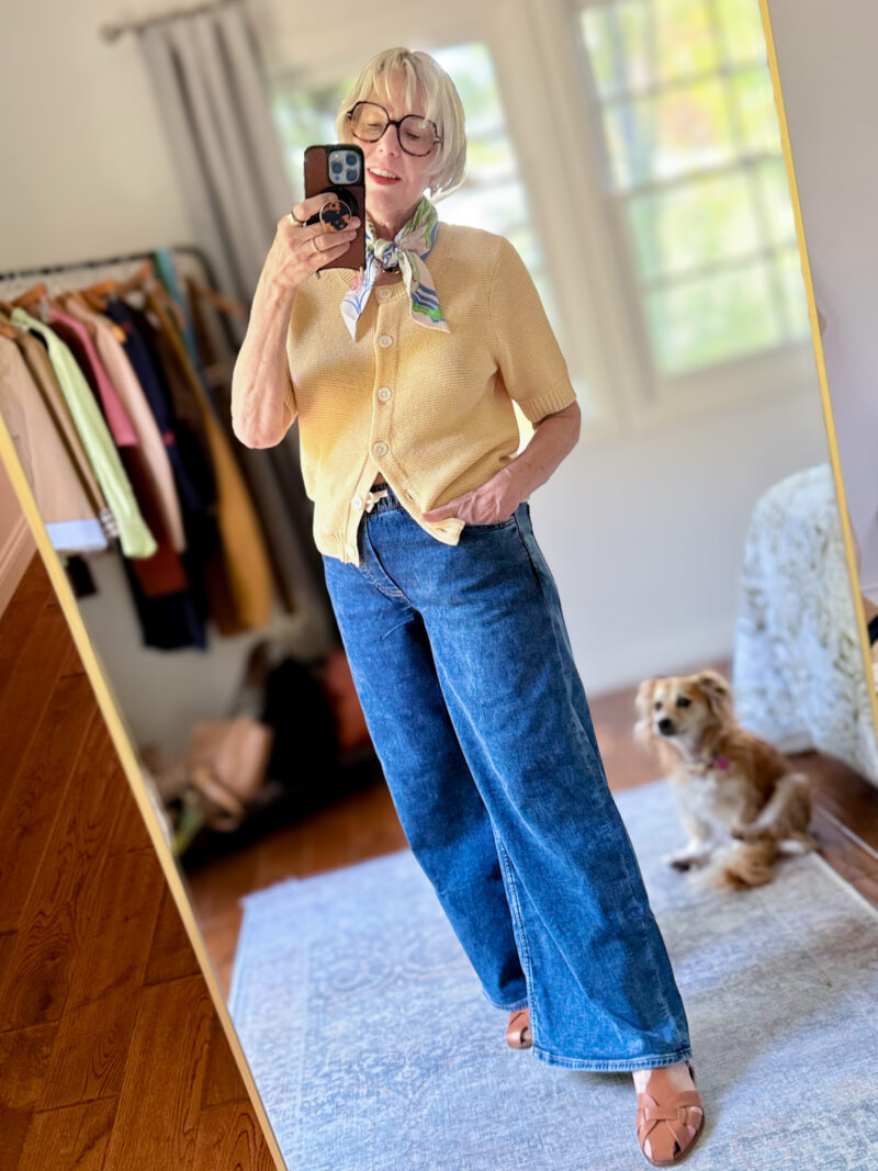 Mirror selfie outfit photo: a yellow short-sleeve knit cardigan buttoned over wide-leg dark wash jeans, styled with a floral-print neck scarf and tan woven mule sandals. A small dog is visible in the background.