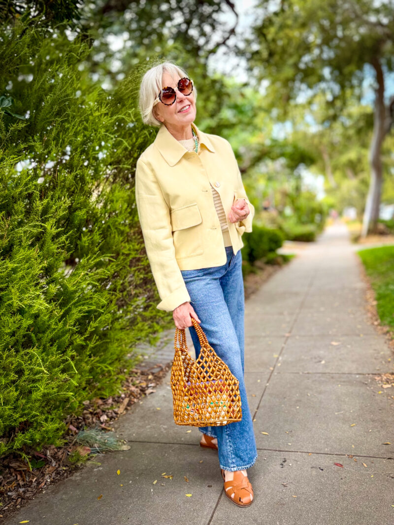 Woman with short silver hair wearing a butter yellow cropped jacket over a striped knit top, medium wash wide-leg jeans, and tan woven leather sandals, carrying a wooden bead bag on a tree-lined sidewalk.