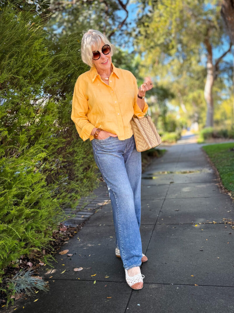 Woman wearing an orange linen shirt with light wash Miramar jeans, white woven flats, and a raffia handbag